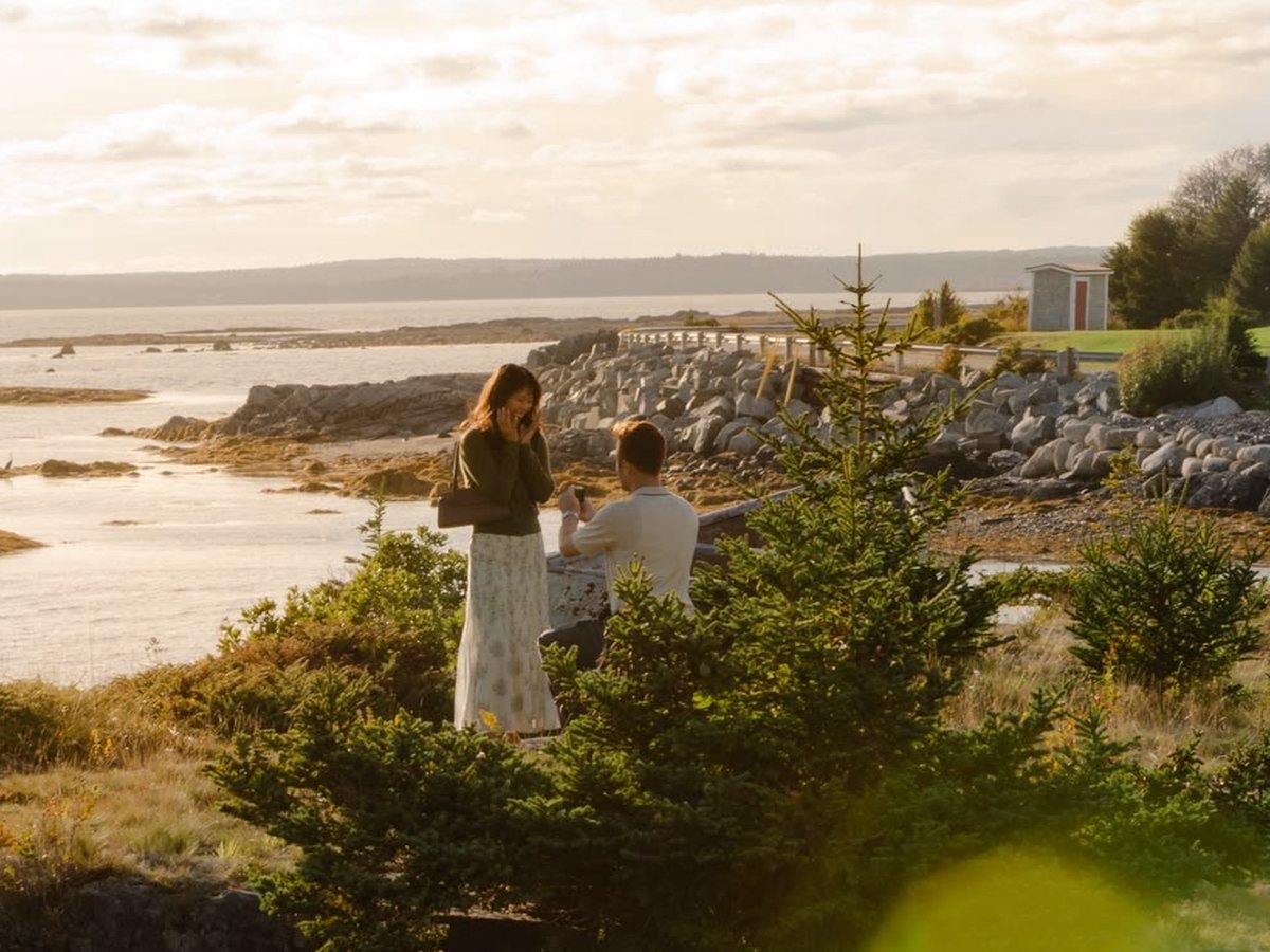 Calin on one knee proposing to Hannah on a rocky shore by the sea, at golden hour.